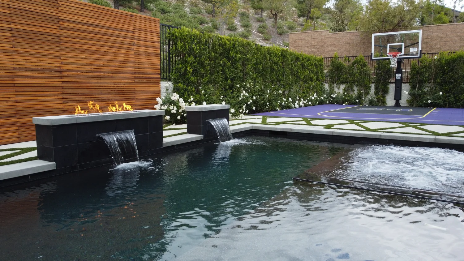 Stunning outdoor backyard with a modern water feature, a basketball hoop, and a beautiful landscaped area with greenery and flowers designed by smart swim pool construction.